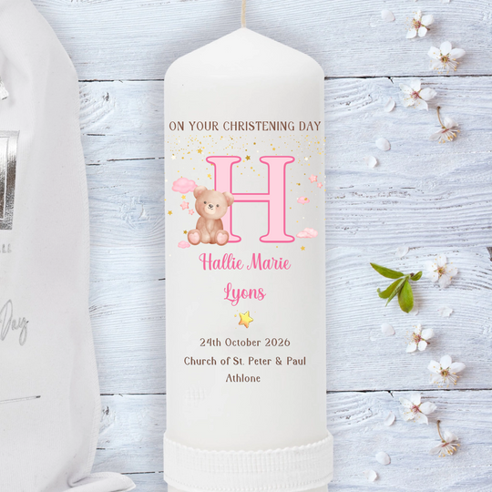 A personalized baby christening candle shown upright on a whitewashed wooden surface, with part of a white garment and small flowers used as styled props. 