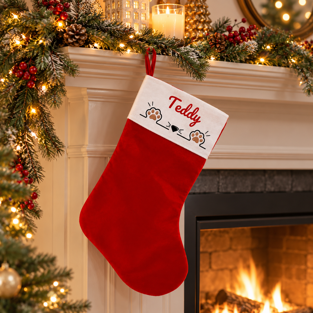 A red Christmas stocking with a white cuff hangs on a fireplace mantel decorated with a lit Christmas tree, pinecones, candles, and red berries. The cuff is personalized with the name 'Teddy' in red script above small paw prints and hearts.