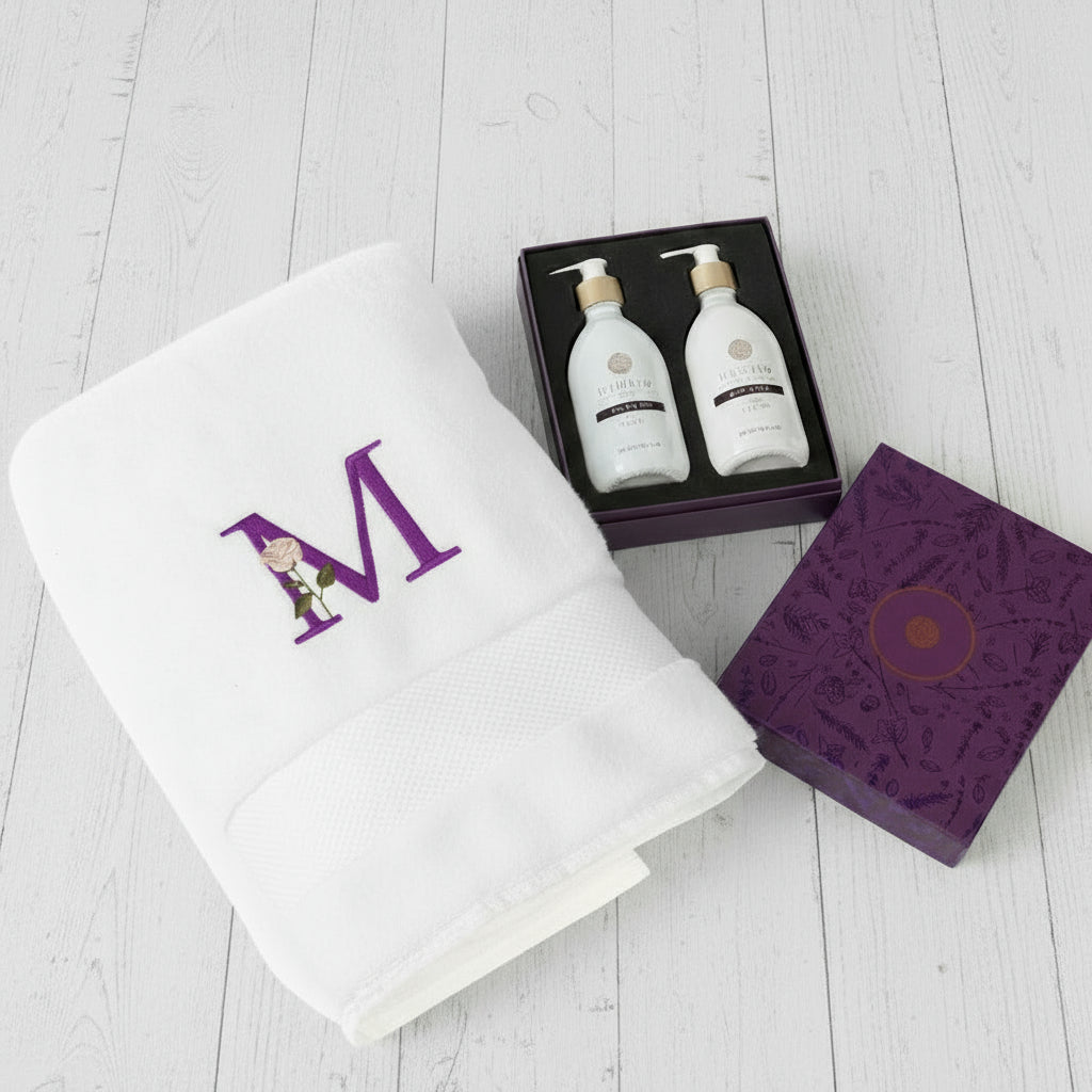 Two white bottles in a purple box with a monogrammed towel and branded box on a white background