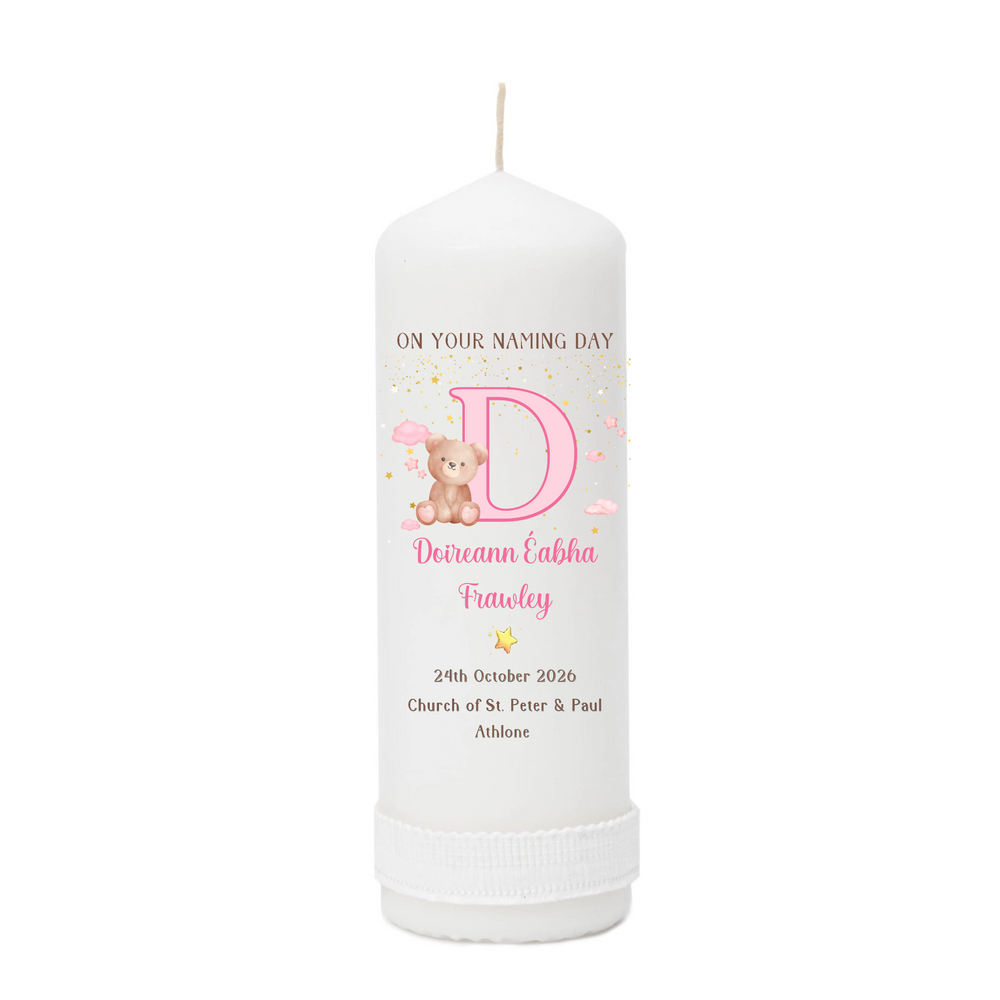 A personalized white pillar candle with a printed pink and gold design featuring a large letter D, a teddy bear, and custom text for a baby naming day.