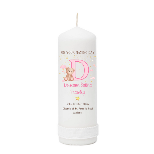 A personalized white pillar candle with a printed pink and gold design featuring a large letter D, a teddy bear, and custom text for a baby naming day.