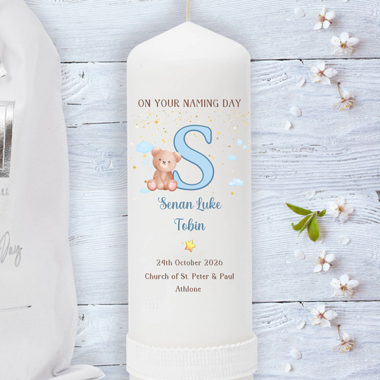 A personalized cream-colored naming day candle with a custom name, date, and initial 'S' printed on it, styled on a white wooden background with small white flowers.