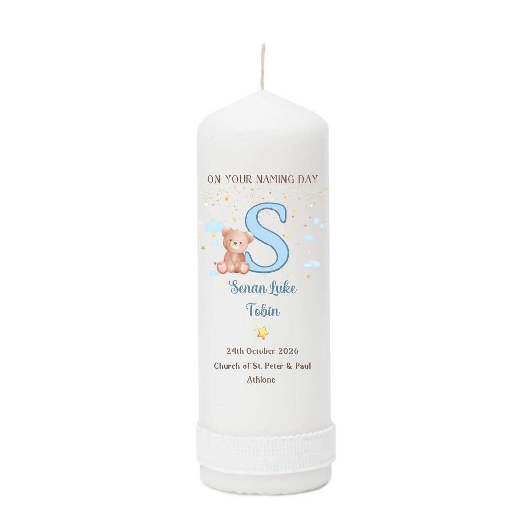 A personalized white pillar candle with a printed name design featuring a large blue letter 'S', a teddy bear, and custom text for a baby naming day.