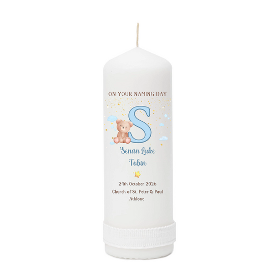 A personalized white pillar candle with a printed name design featuring a large blue letter 'S', a teddy bear, and custom text for a baby naming day.