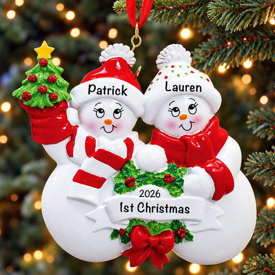 Personalized Christmas snowman ornament hanging on a tree, featuring two snowmen named Patrick and Lauren with red scarves, a small decorated tree, holly, and a banner reading '2026 1st Christmas'.