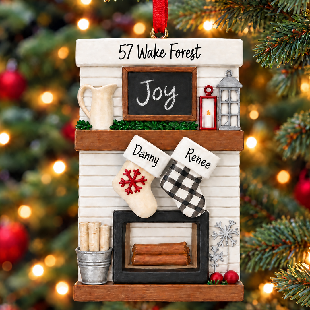A personalized Christmas tree ornament shaped like a fireplace mantel hanging in front of a blurred Christmas tree with warm lights. The ornament includes a white shiplap-style fireplace, a small chalkboard sign reading 'Joy', a red hanging ribbon, two stockings labeled 'Danny' and 'Renee', and holiday decor accents.
