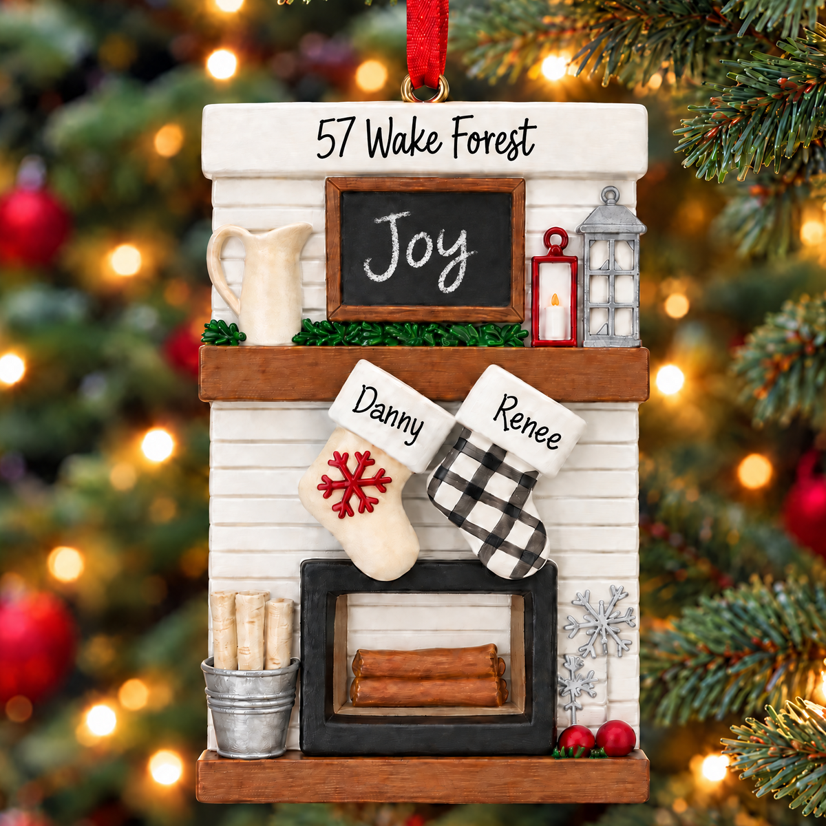 A personalized Christmas tree ornament shaped like a fireplace mantel hanging in front of a blurred Christmas tree with warm lights. The ornament includes a white shiplap-style fireplace, a small chalkboard sign reading 'Joy', a red hanging ribbon, two stockings labeled 'Danny' and 'Renee', and holiday decor accents.