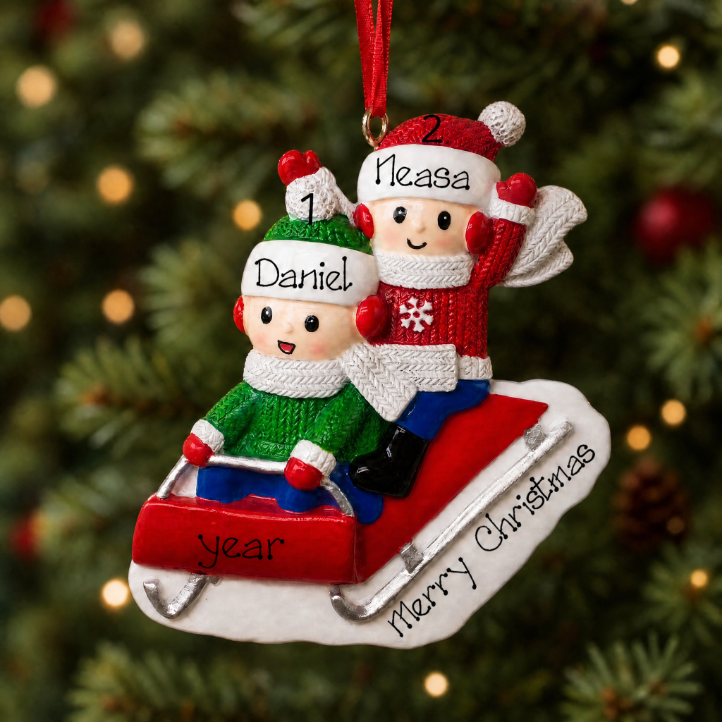 Personalized Christmas ornament featuring two children in holiday sweaters riding a red sled, hanging in front of a blurred Christmas tree background. The names 'Daniel' and 'Neasa' are printed on the children's hats, and 'Merry Christmas' is written on the sled.