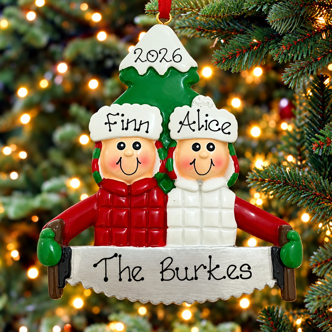 A personalized Christmas ornament shaped like two snowman figures standing together under a green tree topper, with the names Finn and Alice on their hats, the year 2026 at the top, and a banner reading The Burkes at the bottom. The ornament is hanging on a Christmas tree with warm blurred lights in the background.