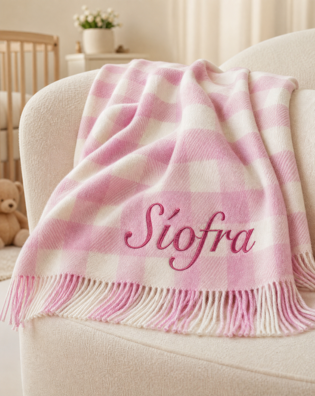 A pink and white checkered throw blanket with fringe and the name 'Siofra' embroidered in pink script, draped over a cream-colored armchair in a softly lit nursery setting.