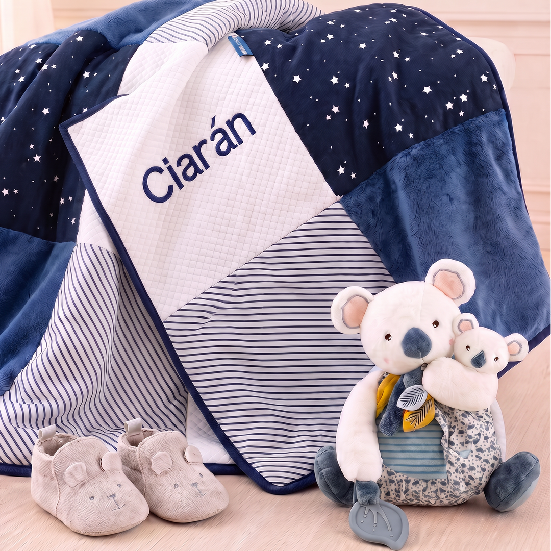 A navy and white patchwork baby blanket with starry and striped sections, personalized with the name 'Ciarán' in navy text on a white panel. The blanket is styled on a light wood floor with a plush koala toy and a pair of baby booties.