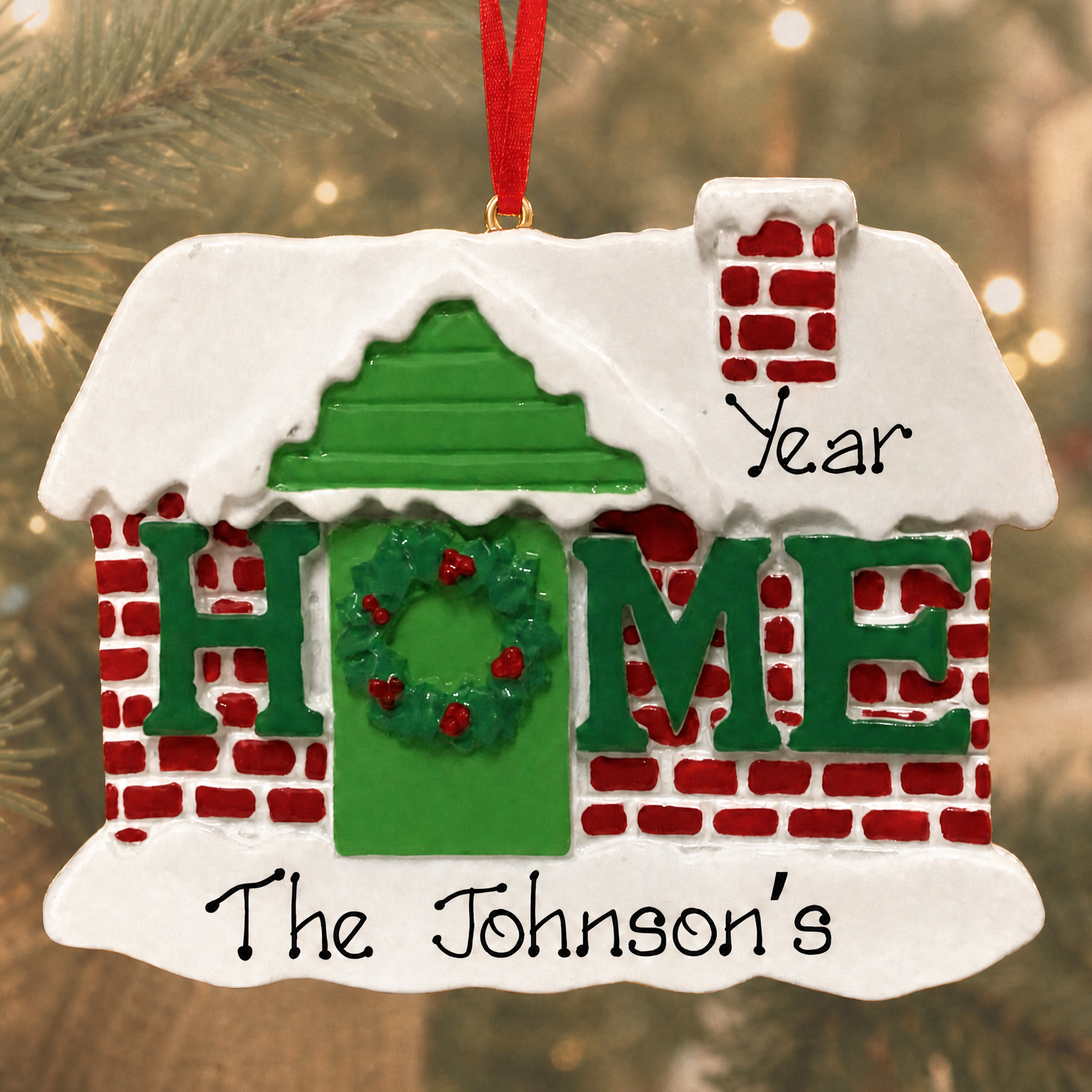 Personalized Christmas house ornament hanging on a Christmas tree, with a red hanging ribbon, white snow-textured roof and base, green door and lettering, and red brick detailing. The ornament is customized with the words 'Year', 'Home', and 'The Johnson's'.