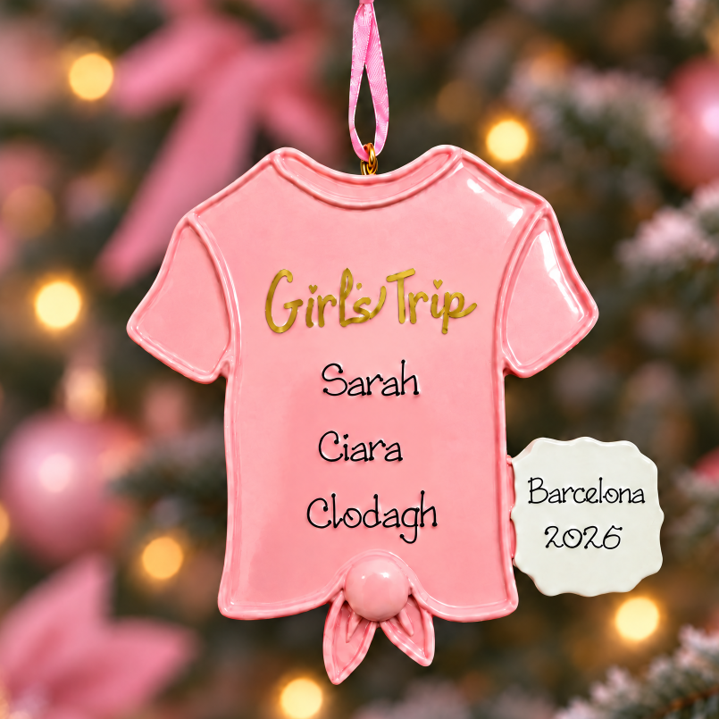 A pink ceramic Christmas ornament shaped like a short-sleeve baby t-shirt hangs from a tree branch. The shirt is printed with the text 'Girls Trip' in gold, followed by customizable names 'Sarah', 'Ciara', 'Clodagh', and 'Barcelona 2026' in black.