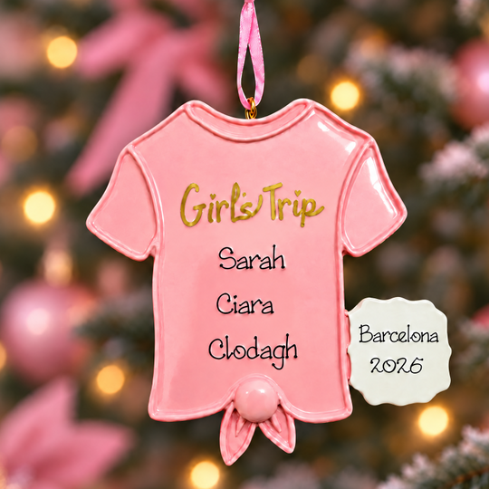 A pink ceramic Christmas ornament shaped like a short-sleeve baby t-shirt hangs from a tree branch. The shirt is printed with the text 'Girls Trip' in gold, followed by customizable names 'Sarah', 'Ciara', 'Clodagh', and 'Barcelona 2026' in black.