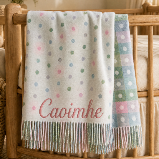 A soft white baby blanket with pastel polka dots and the name 'Caoimhe' in pink script, draped over a wooden crib in a warm, softly lit nursery.