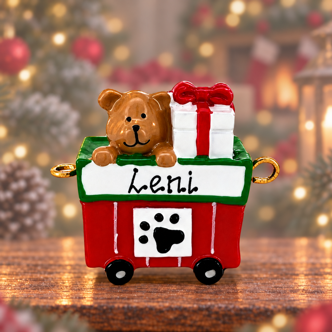 A Christmas ornament shaped like a red and green dog cart with a brown teddy bear sitting inside, next to a white gift box tied with a red bow. The cart has a white front panel with the word 'Lelli' in black script and a black paw print, set on a wooden surface with a blurred holiday background.