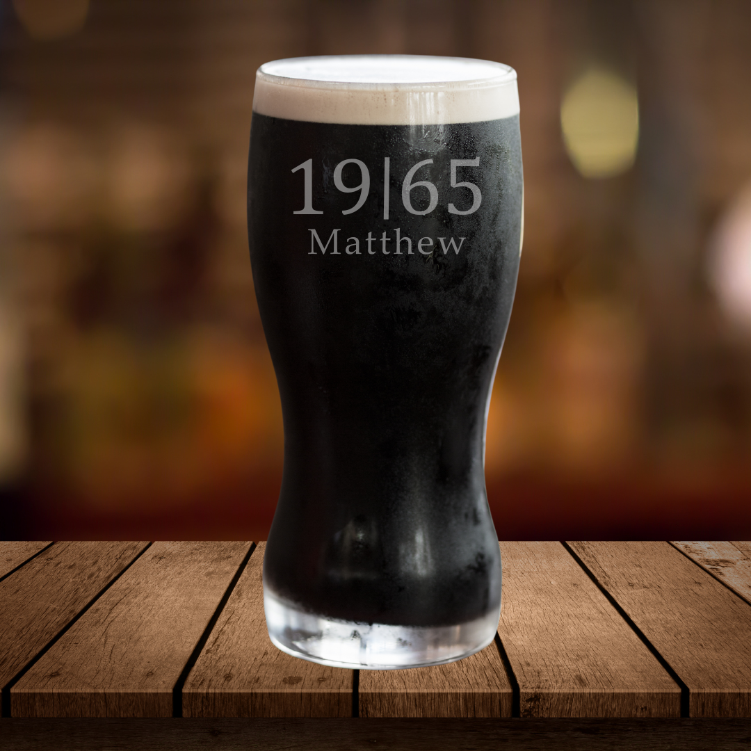 A black pint glass with white foam on top, featuring personalized etched name and year on the side, displayed on a wooden surface with a blurred background.
