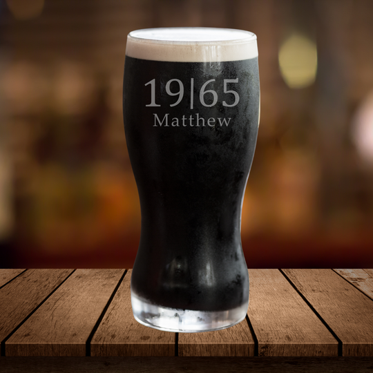 A black pint glass with white foam on top, featuring personalized etched name and year on the side, displayed on a wooden surface with a blurred background.