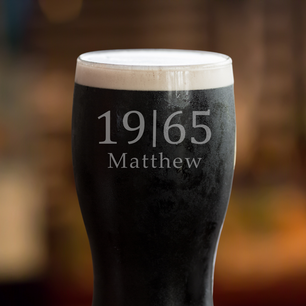 A Guinness pint glass with white foam on top, featuring personalized etched name and year on the side, displayed on a wooden surface with a blurred background.