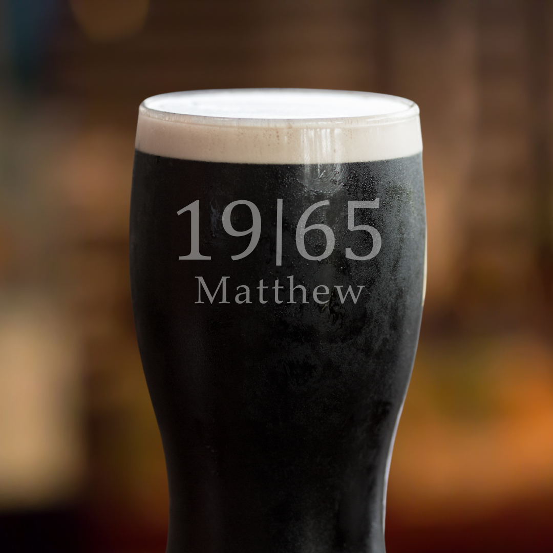 A Guinness pint glass with white foam on top, featuring personalized etched name and year on the side, displayed on a wooden surface with a blurred background.