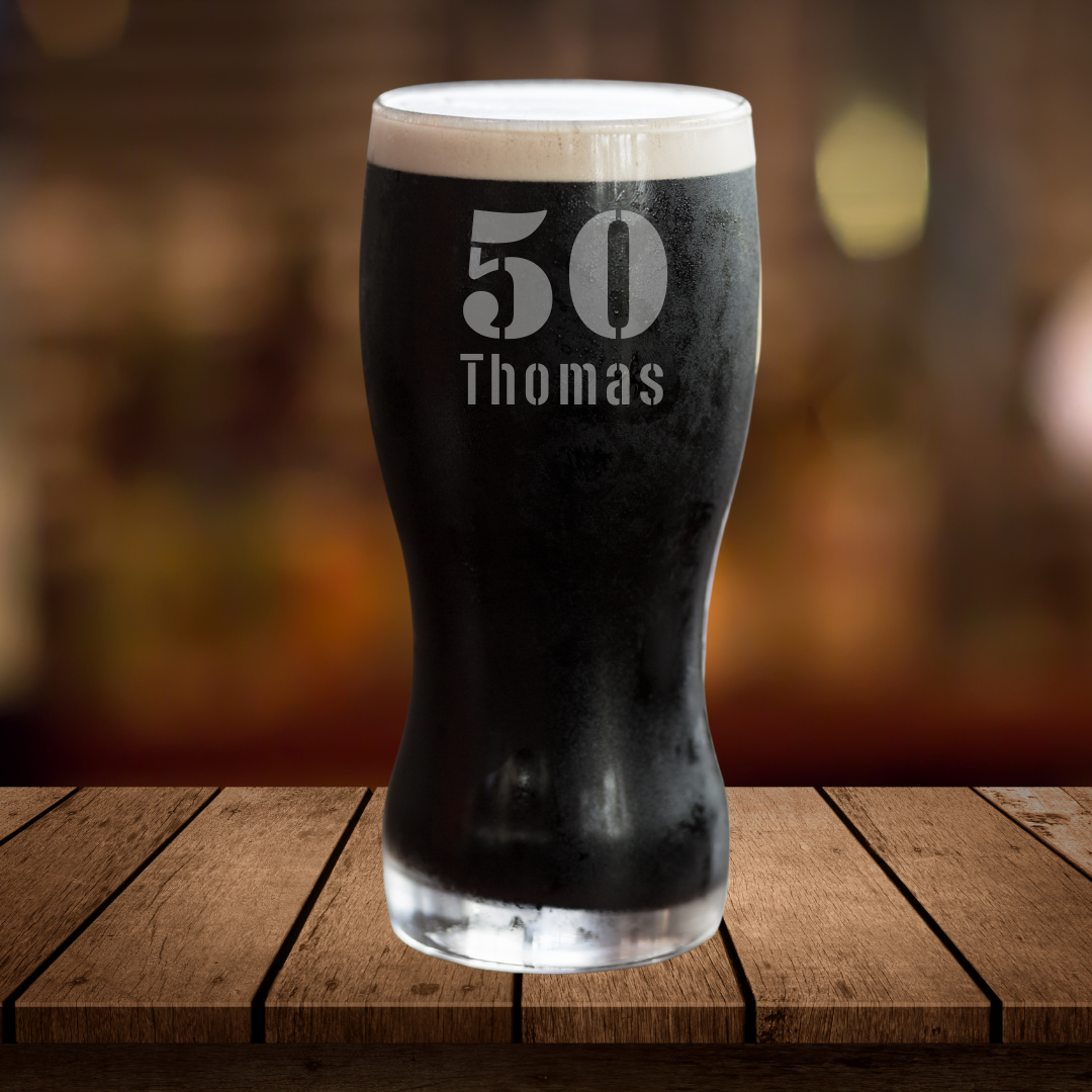 A black pint glass with the number 50 and the name 'Thomas' etched on it, displayed on a wooden surface with a blurred background.