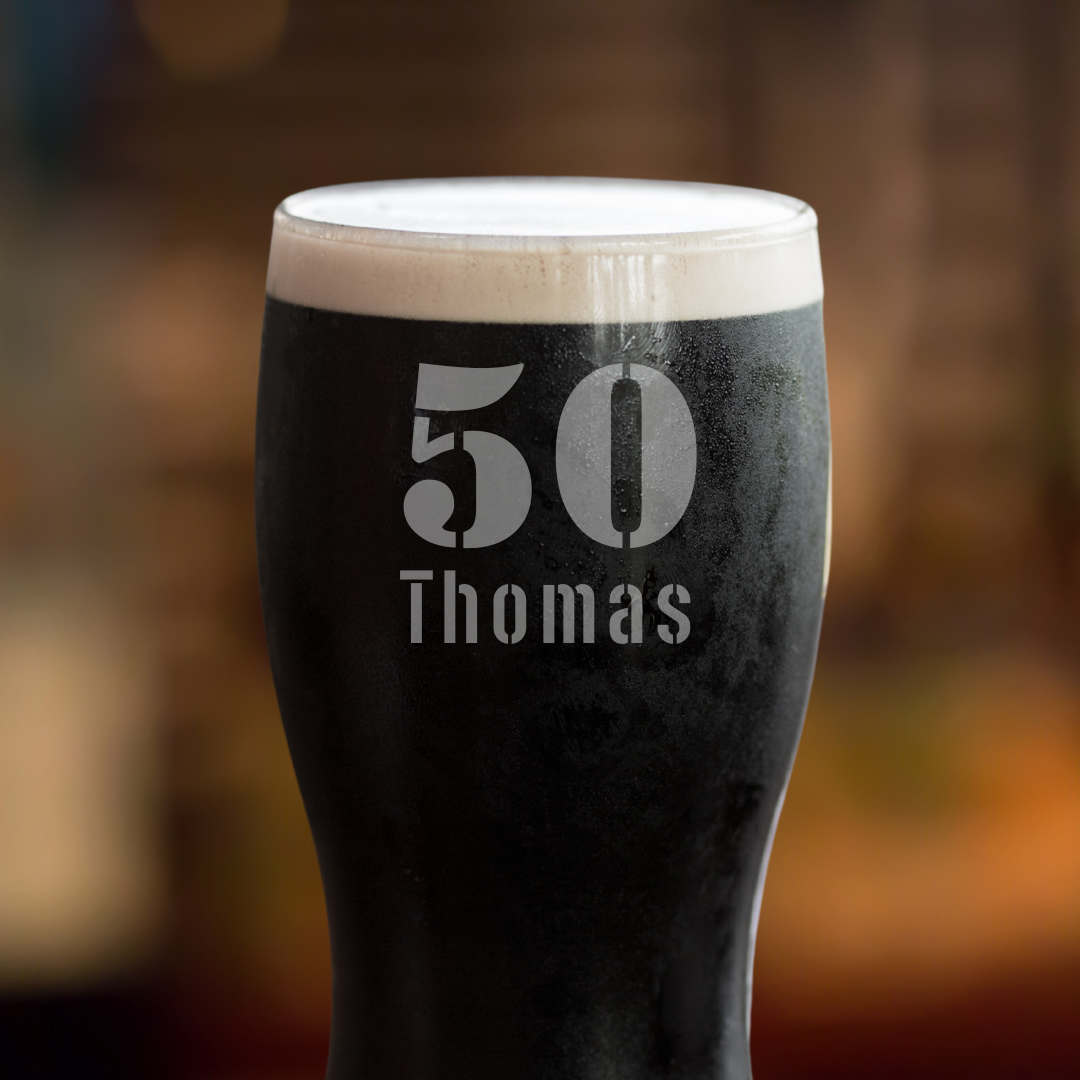 A Guinness pint glass with the number 50 and the name 'Thomas' etched on it, displayed on a wooden surface with a blurred background.