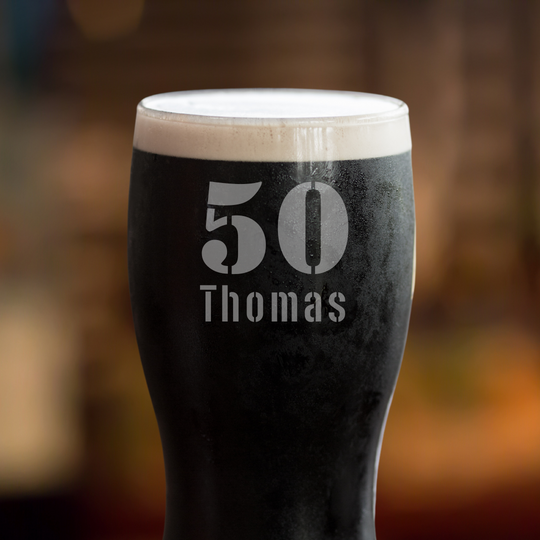 A Guinness pint glass with the number 50 and the name 'Thomas' etched on it, displayed on a wooden surface with a blurred background.