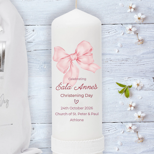 White candle with pink bow design and personalized text on a light wooden background with flowers.