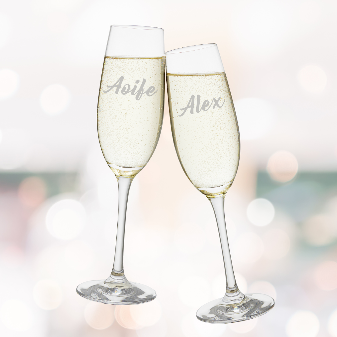 Two clear personalised champagne flutes with etched names, one displaying 'Aoife' and the other 'Alex', against a blurred background.