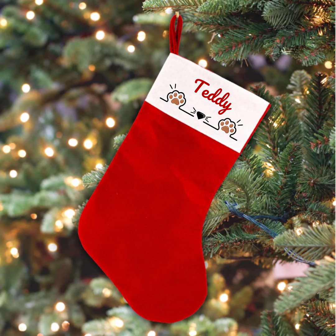 A red Christmas stocking hanging from a decorated Christmas tree. The stocking has a white cuff with the name 'Teddy' written in red script, along with small paw prints and heart illustrations.