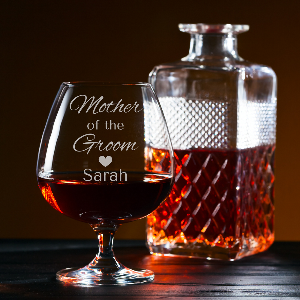 Brandy glass with 'Mother of the Groom Sarah' engraving next to a brandy decanter on a wooden surface.