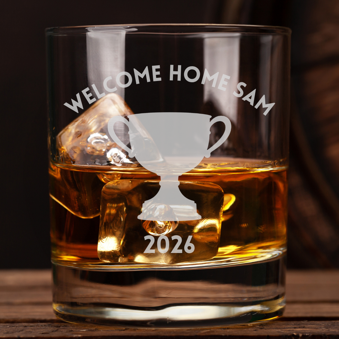 A clear glass rocks tumbler filled with amber whiskey and ice, etched with the words “WELCOME HOME SAM” above a trophy graphic and “2026” below, set on a wooden surface against a dark background.