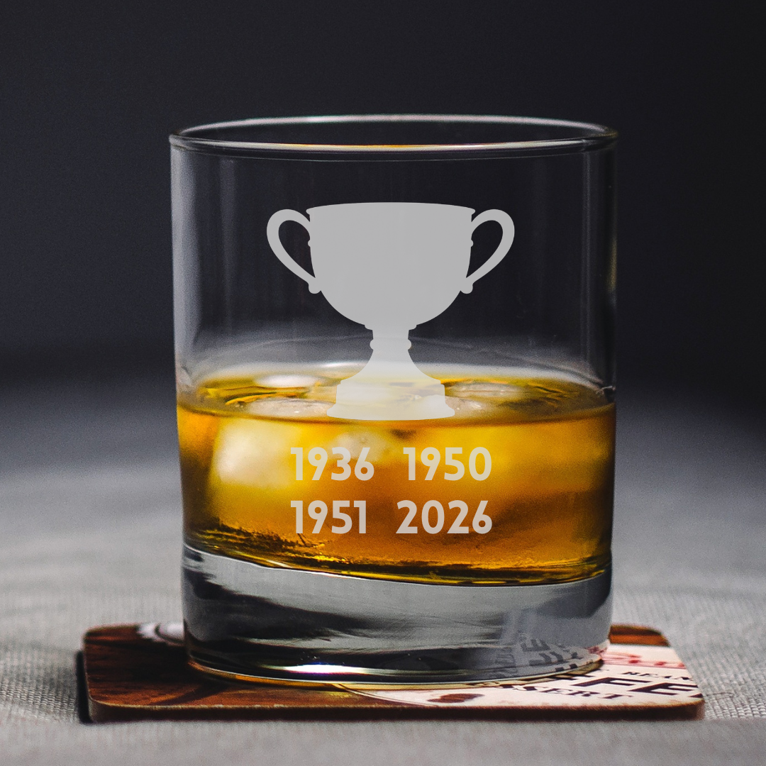 A clear glass rocks tumbler with an etched trophy cup and the years 1936, 1950, 1951, and 2026, filled with amber whiskey and resting on a coaster.