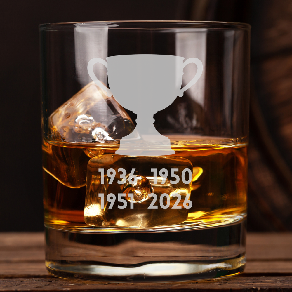 A clear glass rocks tumbler filled with amber whiskey and ice cubes, featuring a frosted white trophy graphic and the years 1936, 1950, 1951, and 2026 etched on the front, set on a dark wooden surface.