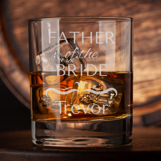 Glass filled with whiskey, engraved with 'Father of the Bride Trevor' on a wooden background.