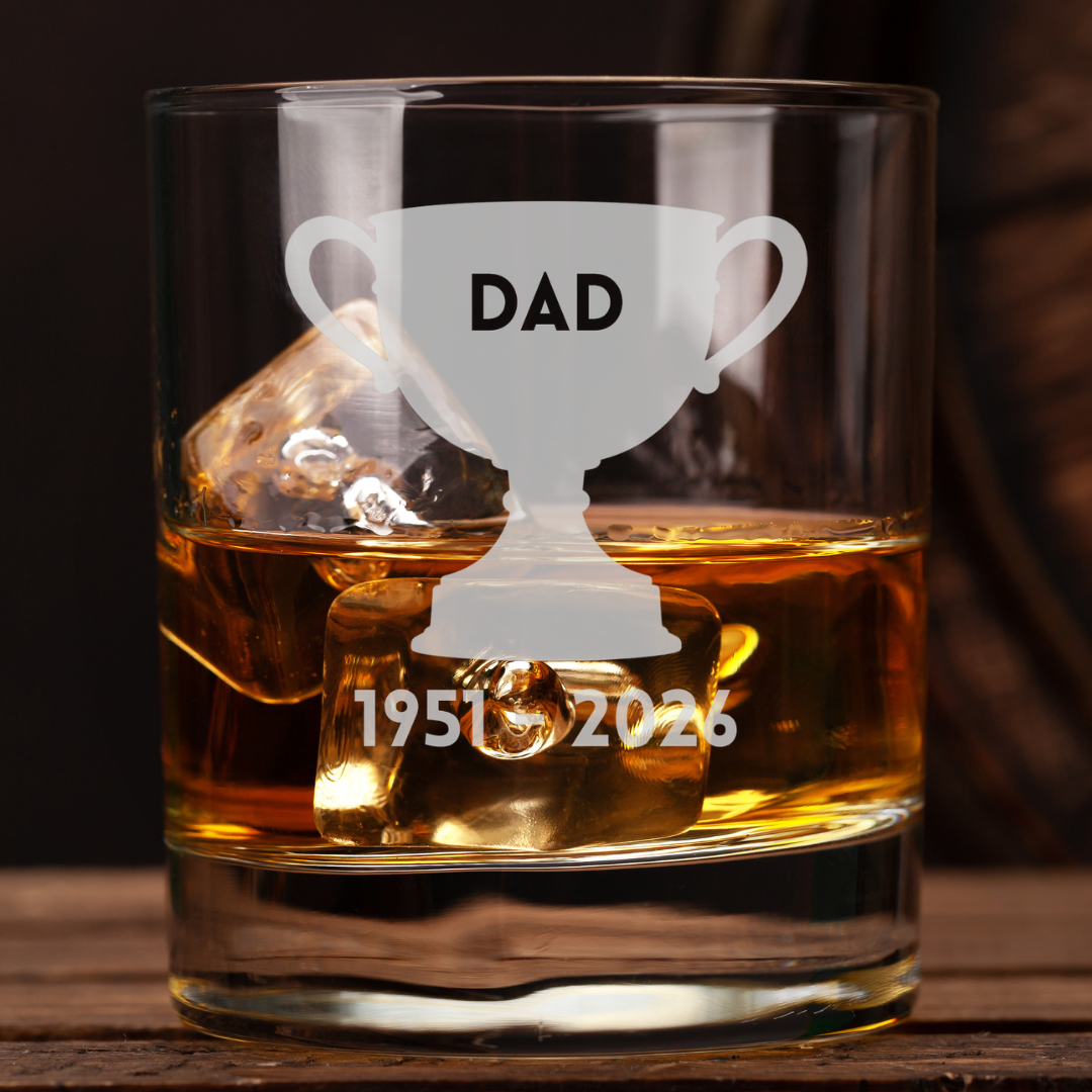 A clear glass rocks tumbler filled with amber whiskey and ice, featuring a frosted white trophy graphic with the word “DAD” and the years “1951-2026” engraved below. The glass sits on a wooden surface against a dark blurred background.