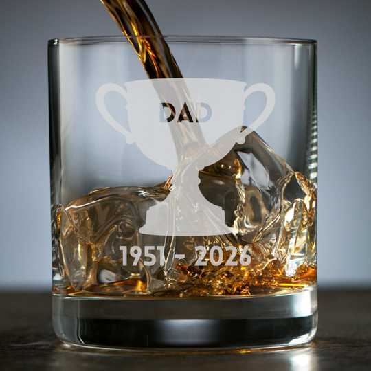 A clear rocks glass with an etched trophy design and the text 'DAD 1951-2026' while amber-colored liquor is being poured into it over ice, set against a dark gray background.