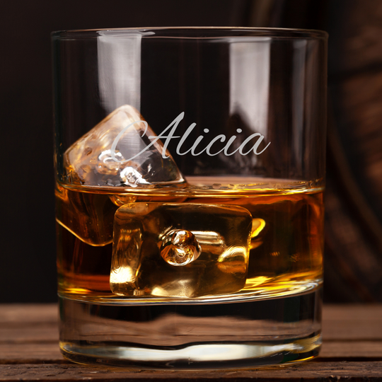 Glass of whiskey with ice cubes and 'Alicia' engraving on a wooden surface.