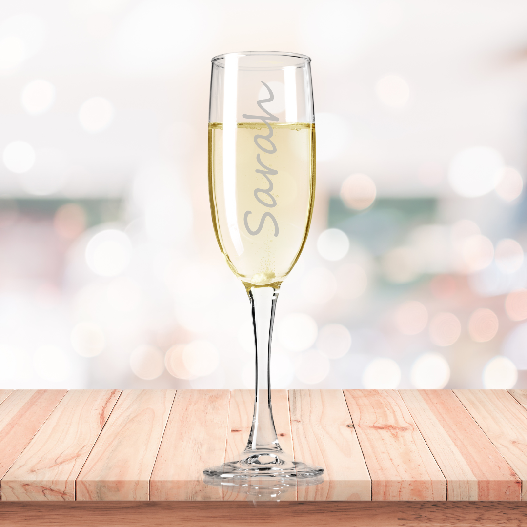 A personalized fine glass champagne flute etched with the name 'Sarah' filled with champagne, presented on a wooden surface with a blurred background.