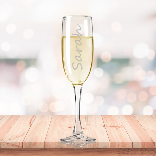 A personalized fine glass champagne flute etched with the name 'Sarah' filled with champagne, presented on a wooden surface with a blurred background.