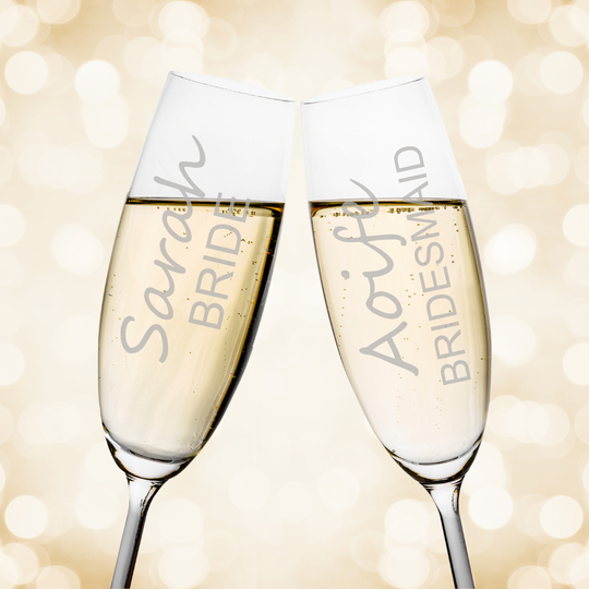Two personalized champagne flutes clinking together, filled with sparkling champagne, against a warm golden bokeh background. One flute is engraved with 'Sarah BRIDE' and the other with 'Aoiife BRIDESMAID'.