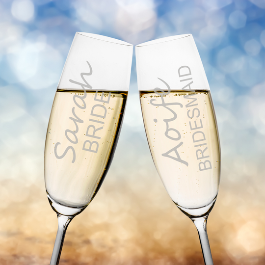 Two personalized champagne flutes clinking together, filled with sparkling champagne, against a soft blue and gold bokeh background. The glasses are engraved with names and titles: 'Sarah BRIDE' and 'Amanda BRIDESMAID'.