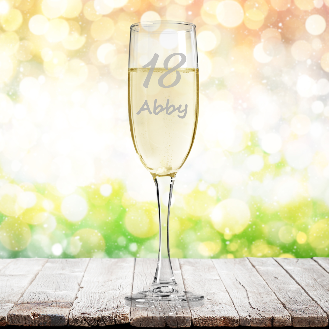 A personalised fine glass champagne flute with the name 'Abby' etched on it, displayed on a wooden surface with a bright blurred background.
