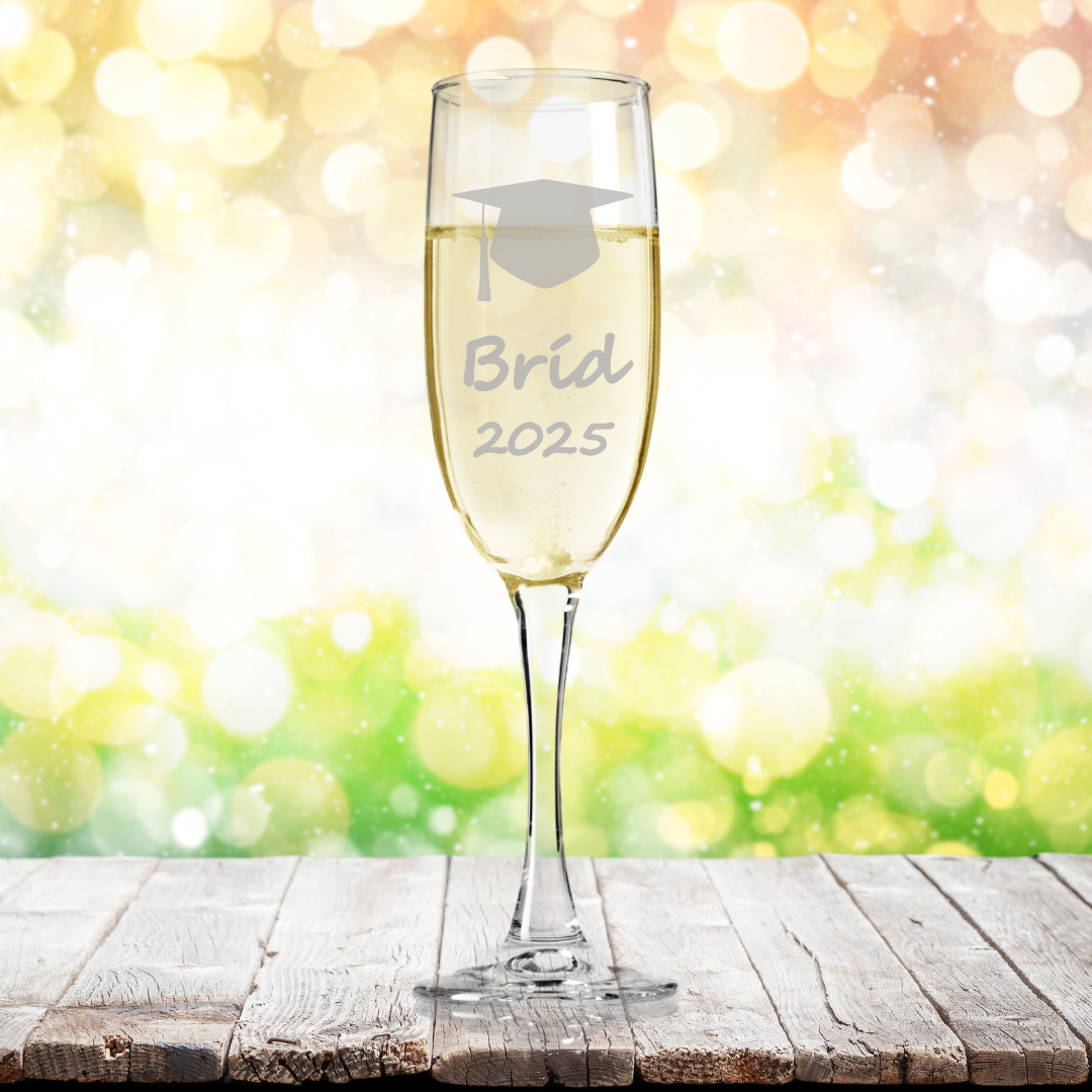 A personalised fine glass champagne flute with a graduation cap clipart, etched name and year 'Brid 2025' on a wooden surface with a blurred bokeh background.