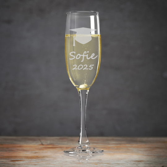 A personalised fine glass champagne flute with a graduation cap clipart, etched name and year 'Brid 2025' on a wooden surface with a grey background.