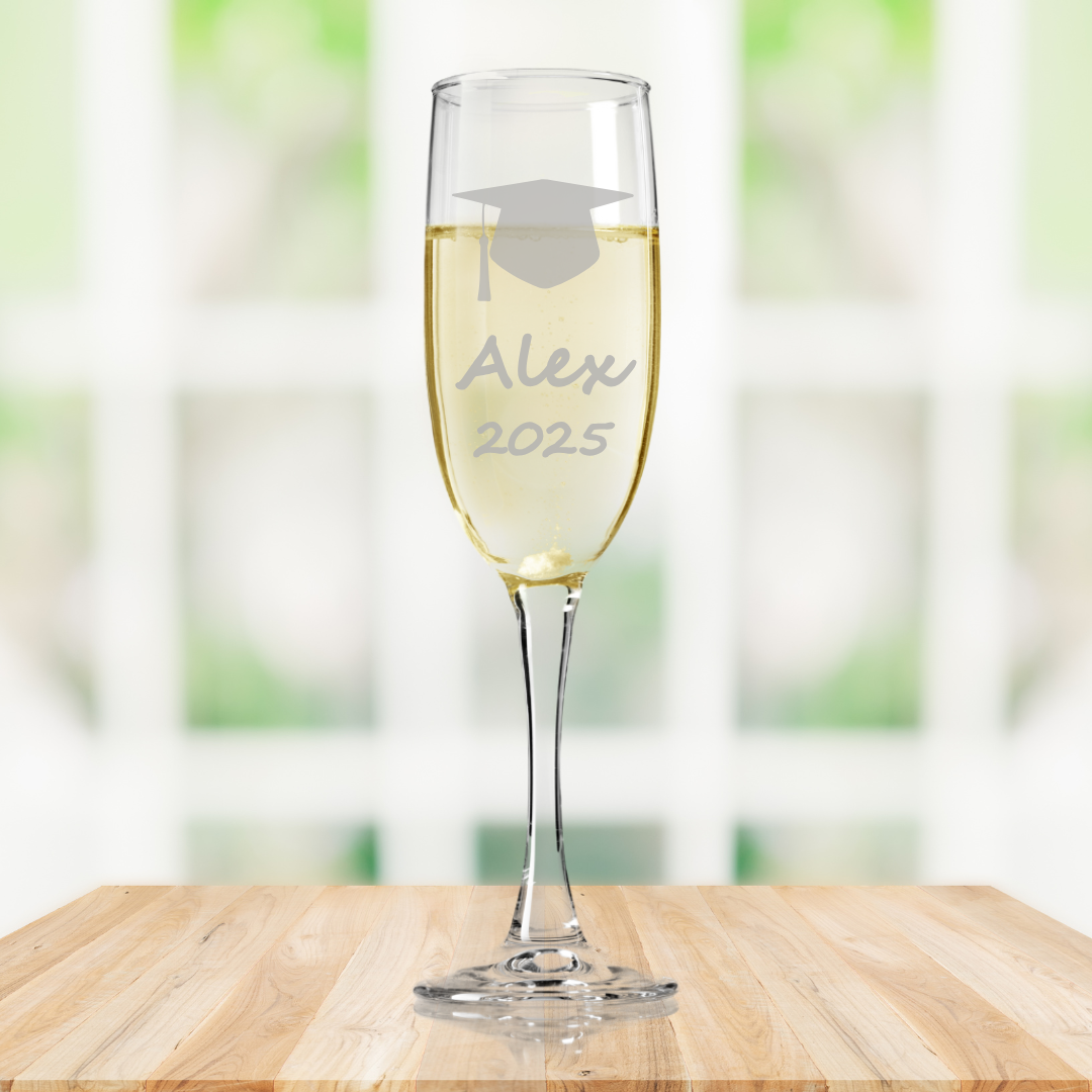 A personalised fine glass champagne flute with a graduation cap clipart, etched name and year 'Brid 2025' on a wooden surface with a blurred green background.