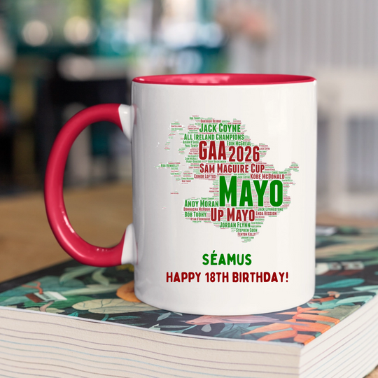 A white ceramic mug with a red interior and red handle sits on top of a colorful book. The mug features a green and red graphic shaped like the map of County Mayo, filled with text celebrating GAA 2026, Sam Maguire Cup, and Mayo, with the name Séamus and birthday message printed below.