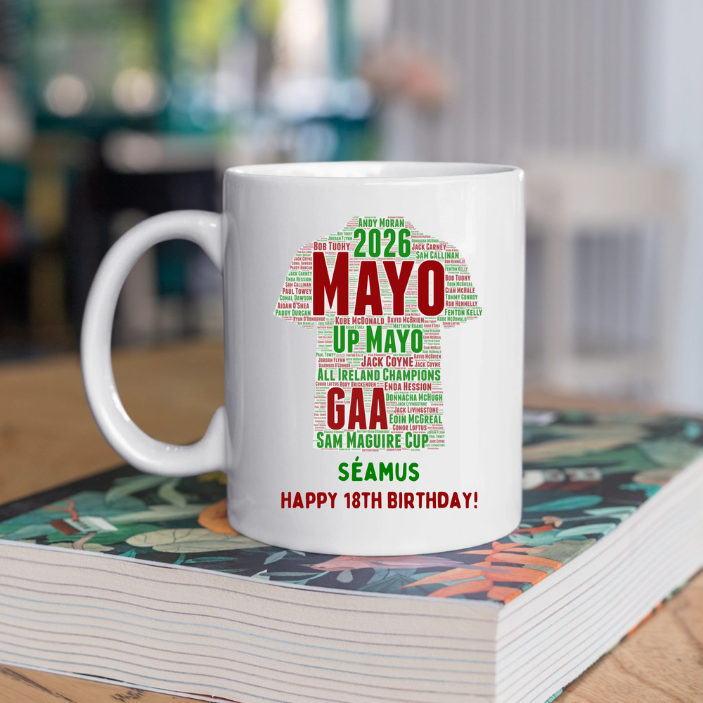 A white ceramic mug with a colorful Mayo jersey text graphic celebrating Sam Maguire's 2026 career, placed on top of a colorful book in a blurred indoor setting.