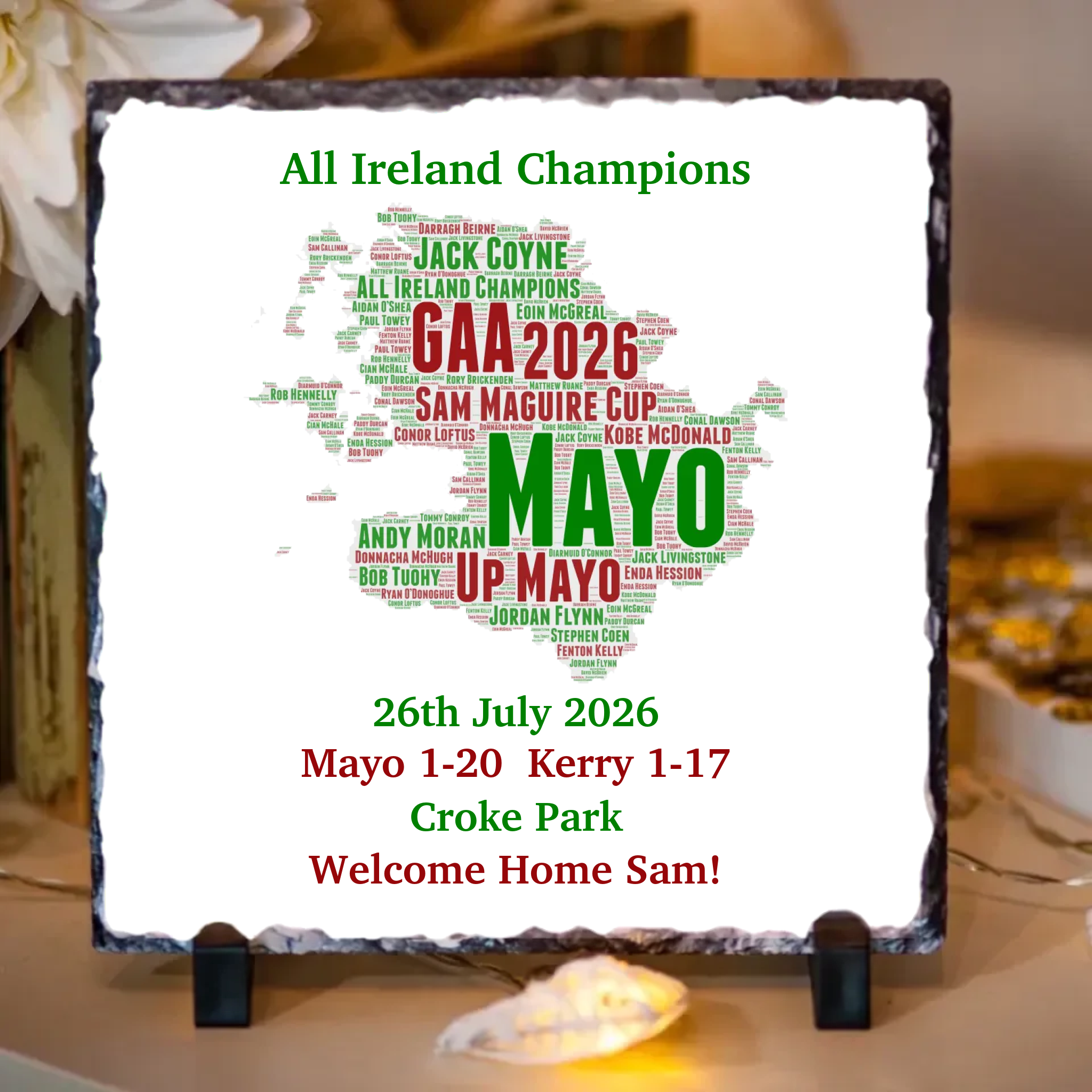 A square slate sign on a small black stand, featuring a white background with a map of Ireland filled with green and red text celebrating GAA winners and match results. The sign reads 'All Ireland Champions' and 'GAA 2026 SAM MAGUIRE CUP MAYO UP MAYO 26th July 2026 Mayo 1-20 Kerry 1-17 Croke Park Welcome Home Sam!'.