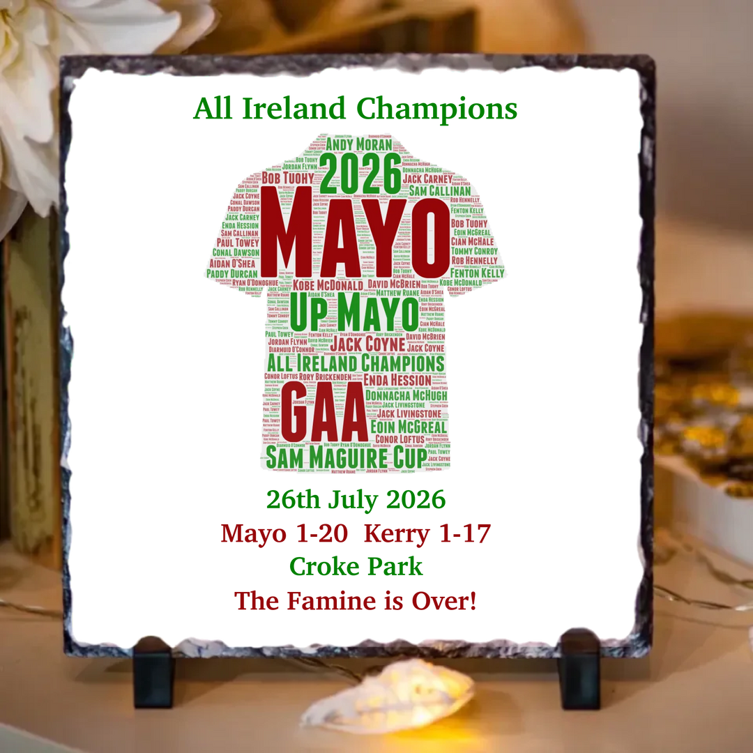 A square decorative sign on a small black stand, featuring a white background with a green and red text design celebrating Mayo GAA and the 2026 Sam Maguire Cup. The sign has a rough dark border and is photographed indoors on a tabletop with blurred decor in the background.